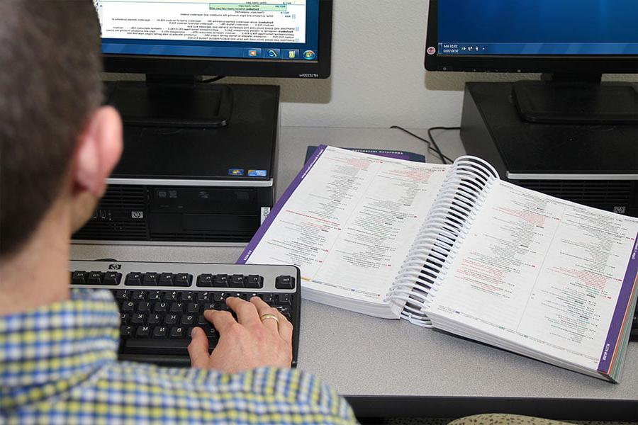 An individual sits at a computer, typing on the keyboard, with a book of medical coding information open on the desk next to them
